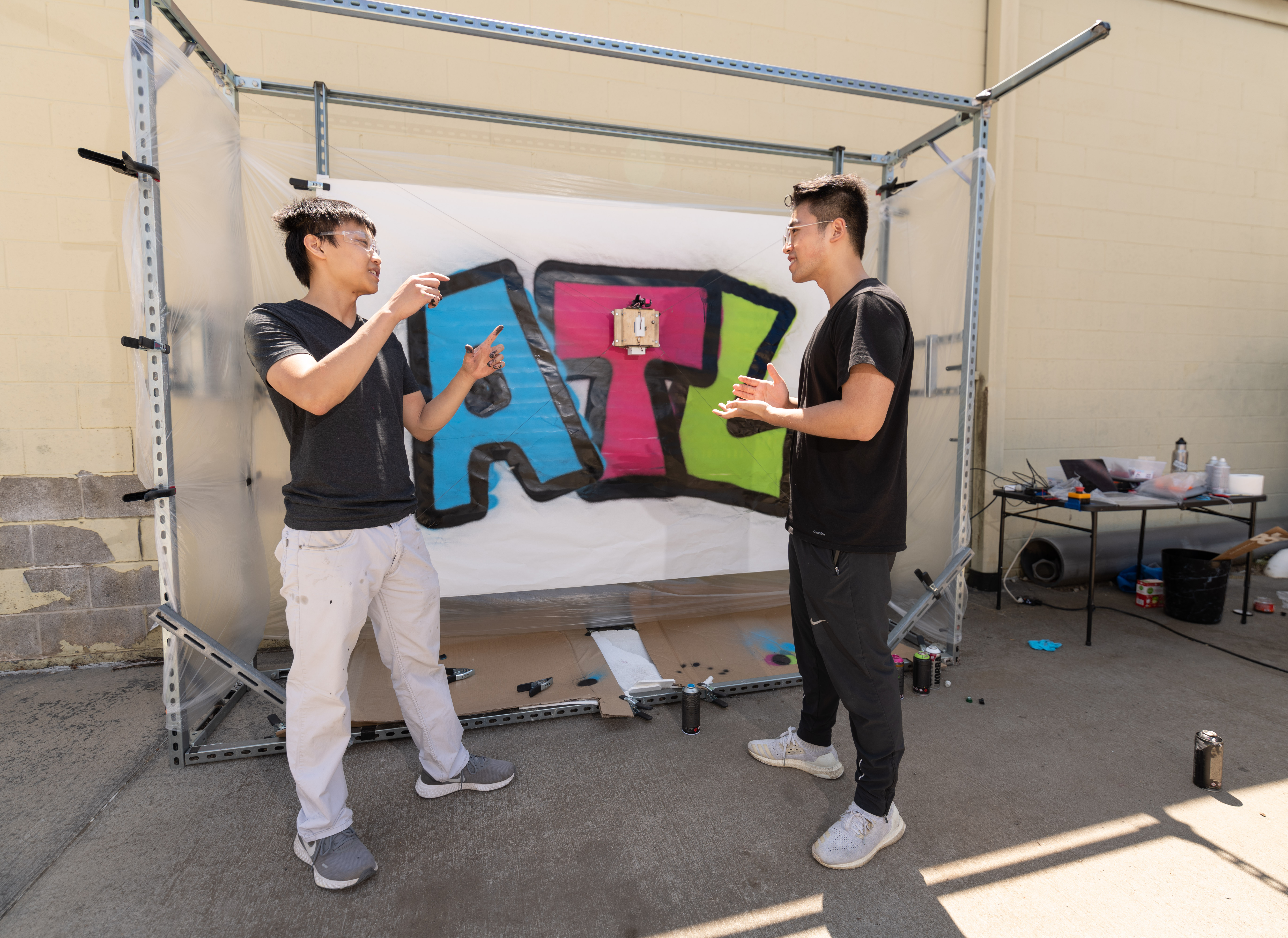Two young men on either side of a canvas where the GTGraffiti robot has painted ATL in blue, pink, and green block letters.