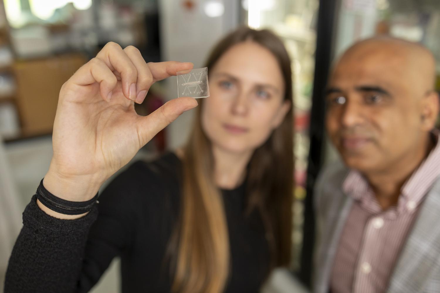 Person holding a small transparent microchip between fingers, with another individual standing nearby in a laboratory setting.