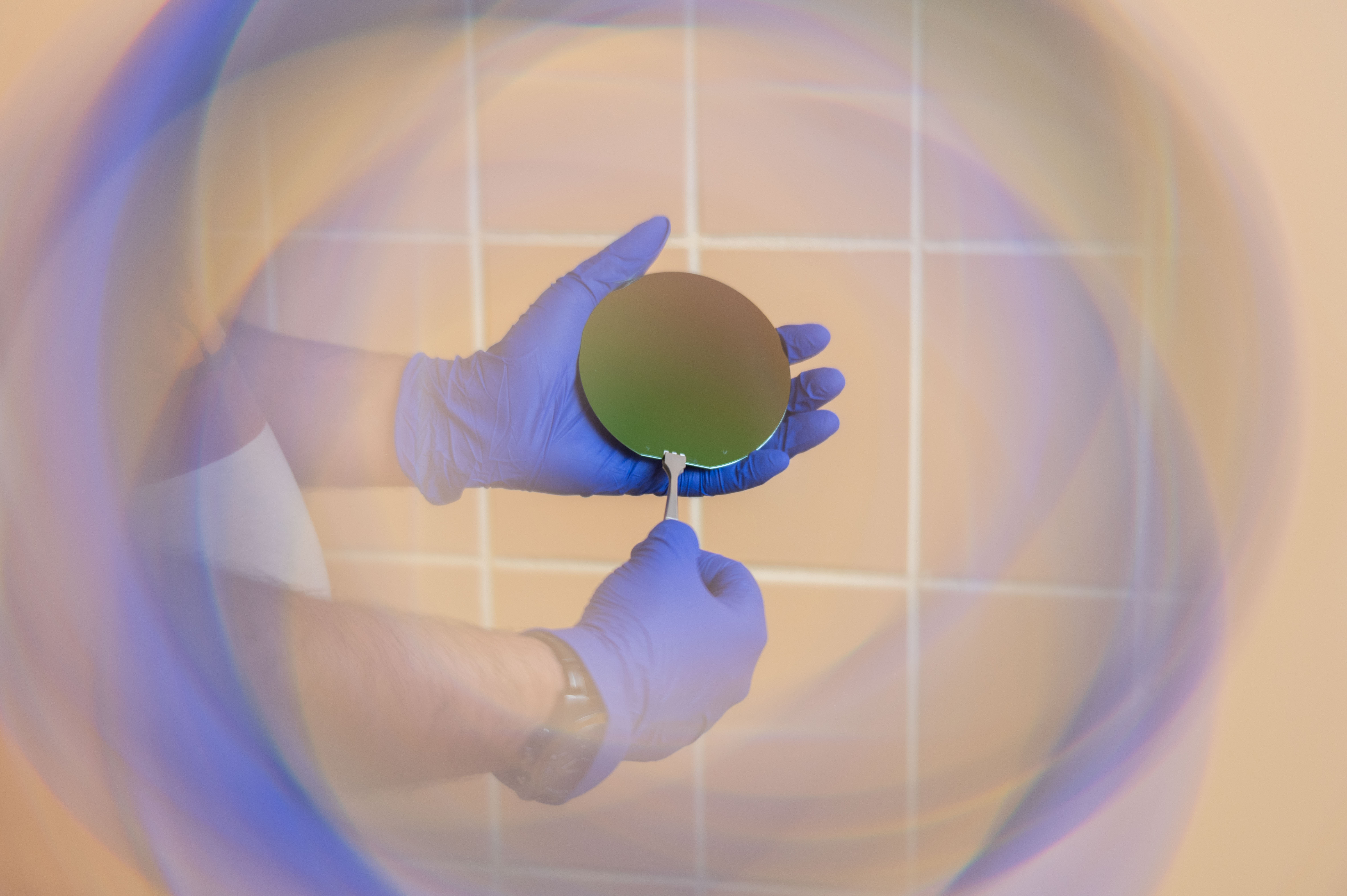 Hands wearing blue gloves holding a circular silicon wafer with a tool, against a tiled background with a colorful blurred ring effect.