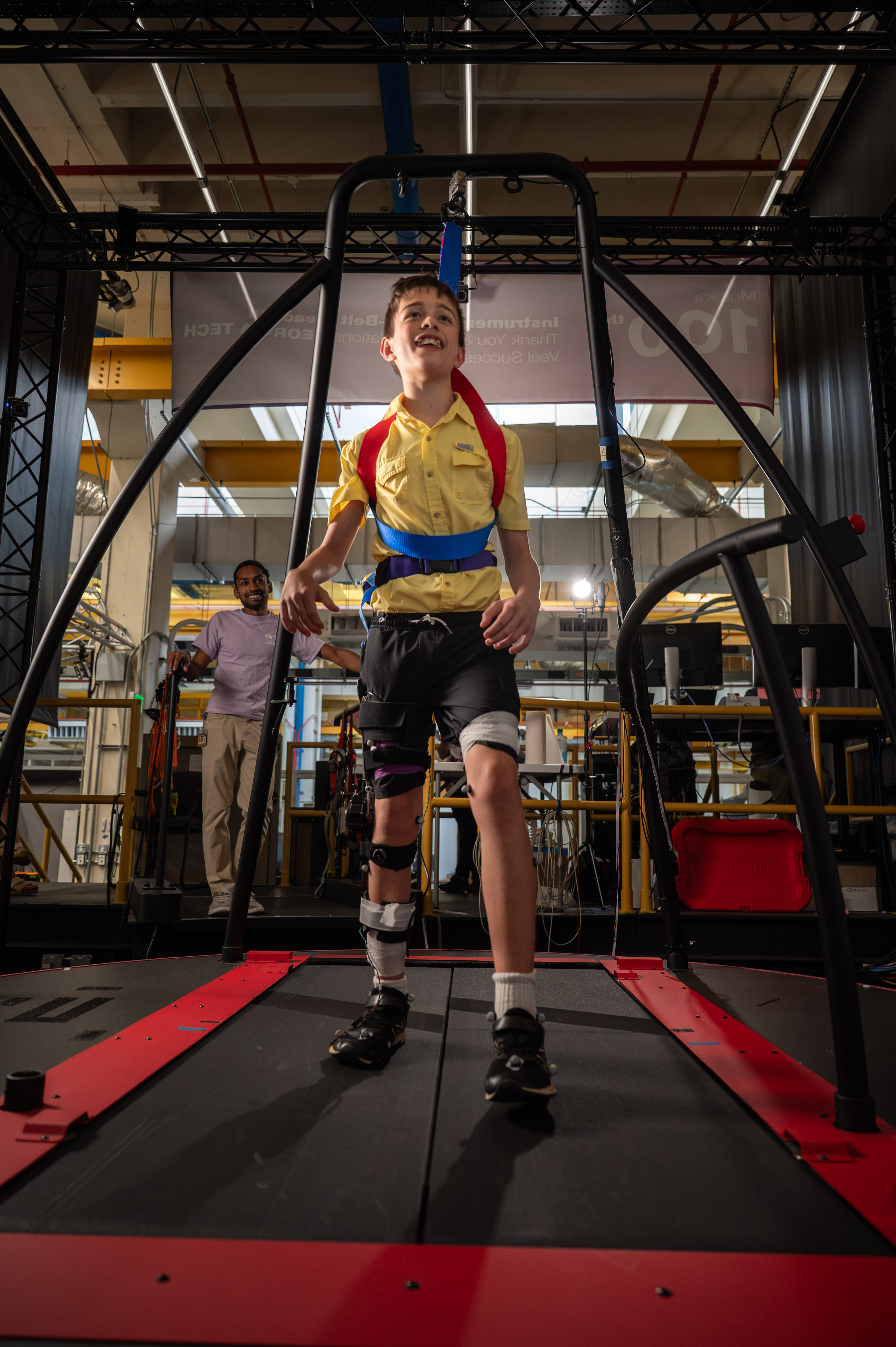 Person wearing a yellow shirt and harness walking on a treadmill inside a robotic gait training system, with sensors and support straps attached to the legs.