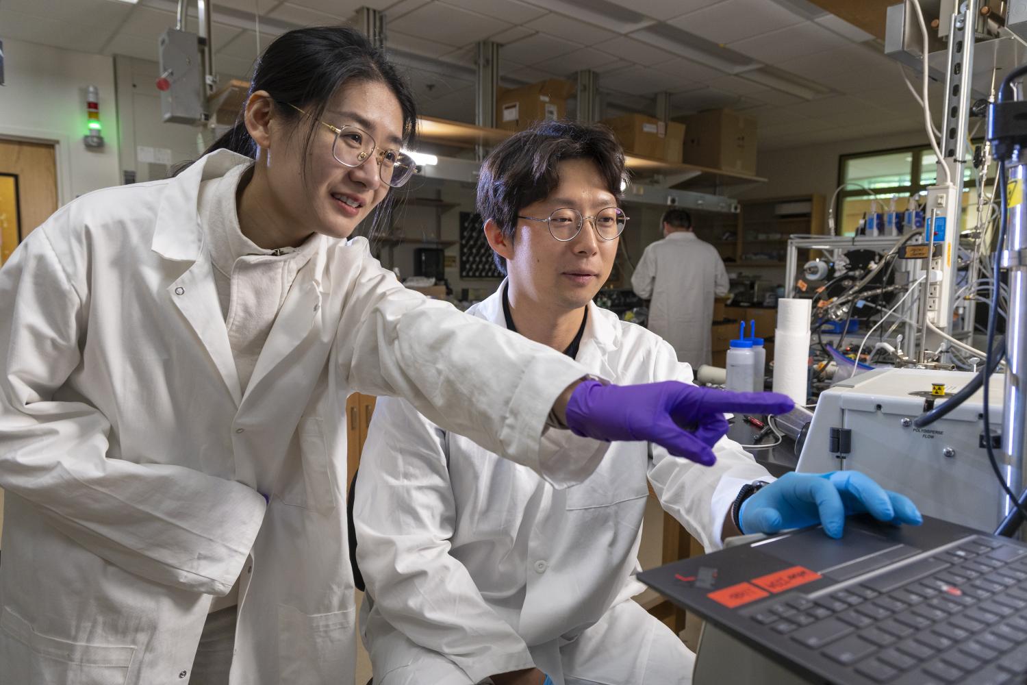 Two researchers in white lab coats working in a laboratory, with one pointing at a laptop screen while the other operates equipment. Various scientific instruments and supplies are visible on the workbench in the background.