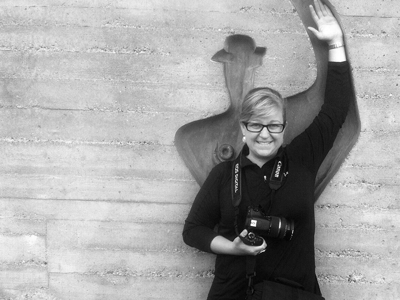 A woman holding a camera stands in front of a relief carving in a concrete wall mimicking its posture, with her arm raised over her head.