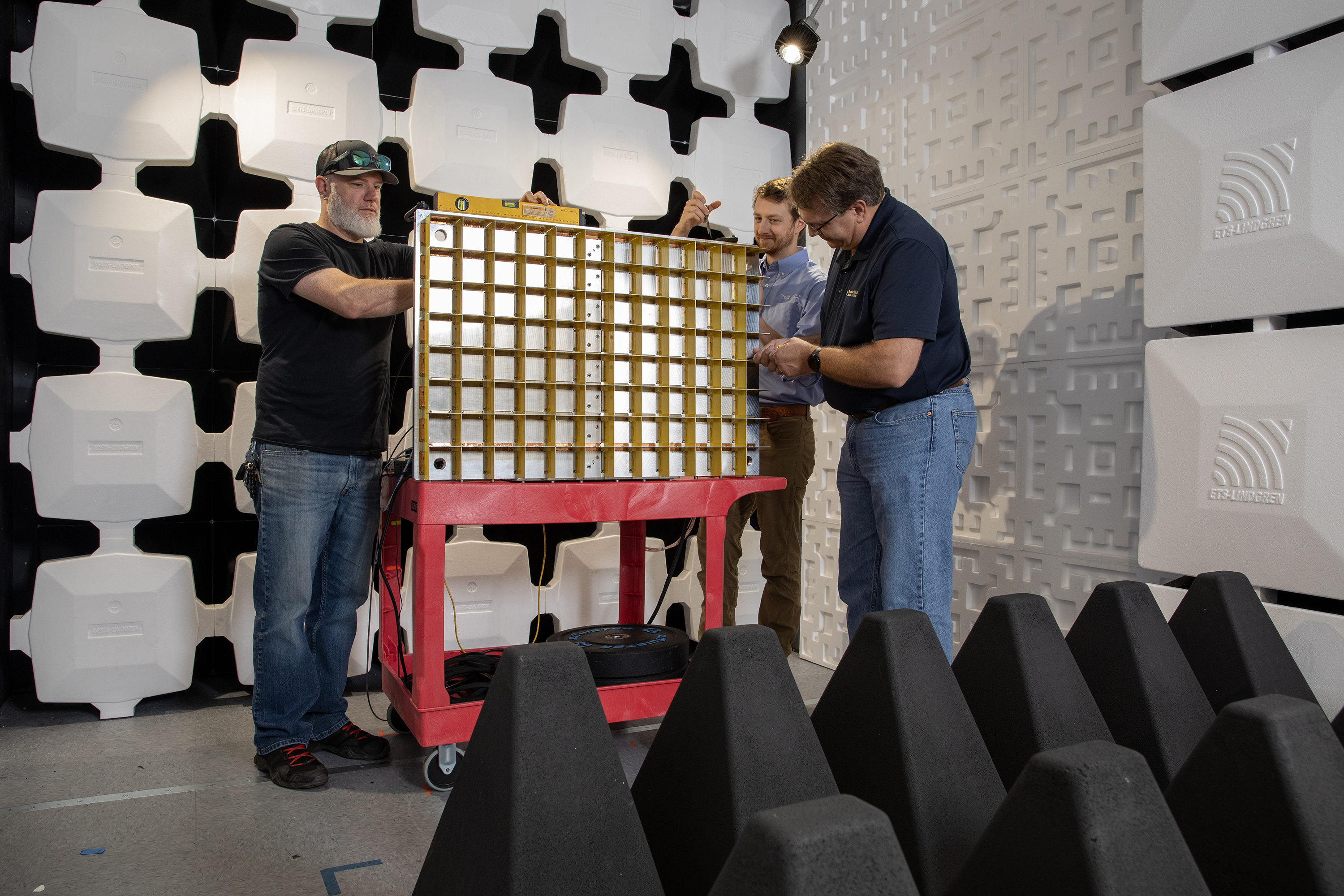 Three people working with a large grid-like metal structure mounted on a red cart inside an acoustic testing room with white soundproof panels and black foam wedges.