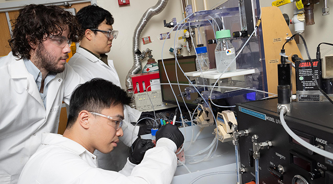 Three men in lab coats working at a bench on an experimental setup with tubes, vials, and pumps.