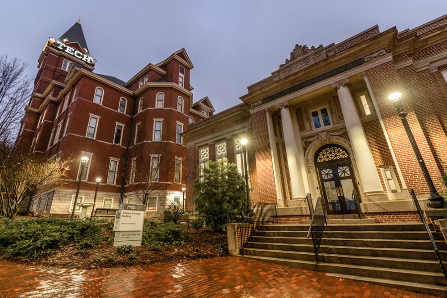 Historic Georgia Tech brick buildings at dusk, including a tower labeled “TECH,” with lit entryway, columns, and wet brick walkway after rain.