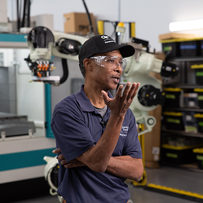 Person wearing a dark polo shirt and a black cap standing in an industrial workspace with robotic machinery and equipment in the background.