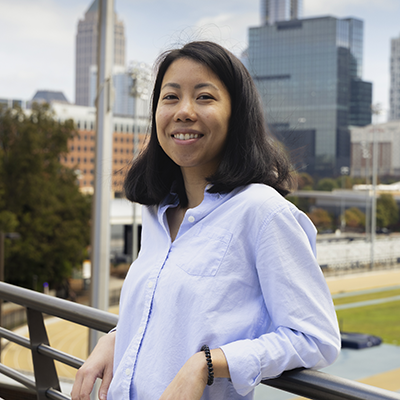 Person wearing a light blue button-up shirt standing outdoors on a balcony with a city skyline in the background, featuring tall modern buildings and trees.