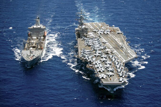 Two large naval vessels sailing side by side in open ocean, including an aircraft carrier with numerous fighter jets on its deck and a support ship nearby.