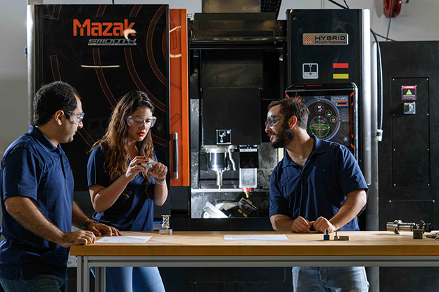 Three individuals standing at a workbench in front of a large Mazak hybrid manufacturing machine, discussing components and technical drawings.
