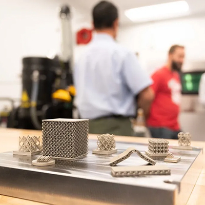 Metal 3D-printed lattice structures and geometric components displayed on a metal plate in a manufacturing lab, with industrial equipment visible in the background.