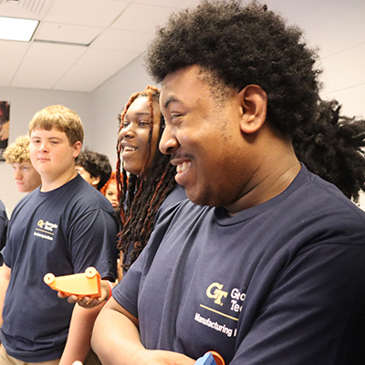 Group of students in navy shirts standing in a classroom, each holding small orange and blue 3D-printed objects.