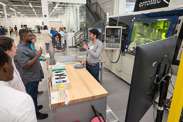 Demonstration in a manufacturing lab where a presenter explains equipment to a small group gathered around a worktable, with industrial machinery in the background.