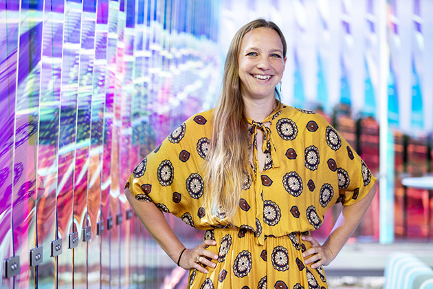 Person in a yellow patterned dress with hands on hips, standing in front of a vibrant, colorful wall with reflective panels