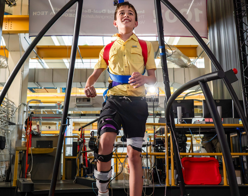  young boy walking with assistance from a structure and a cable holding him up 