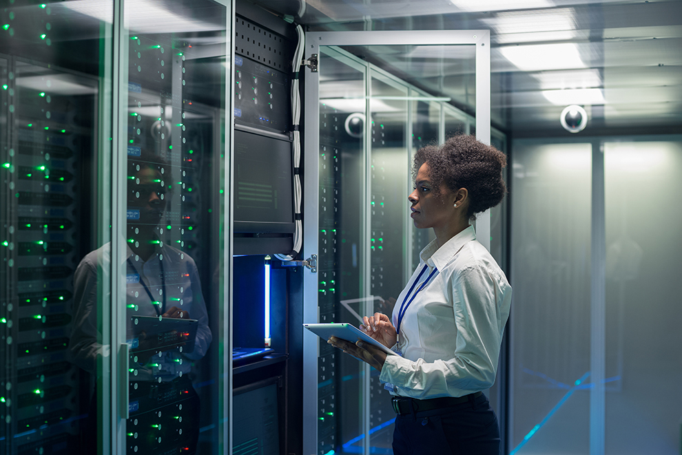 IT professional monitors server racks in a data center, using a tablet to manage systems and network operations.