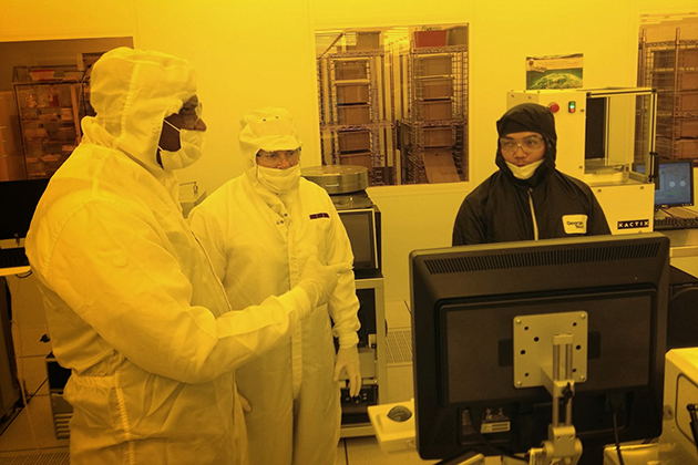 Three people in full cleanroom suits stand around a computer monitor in a yellow‑lit lab, with one person gesturing toward the screen.