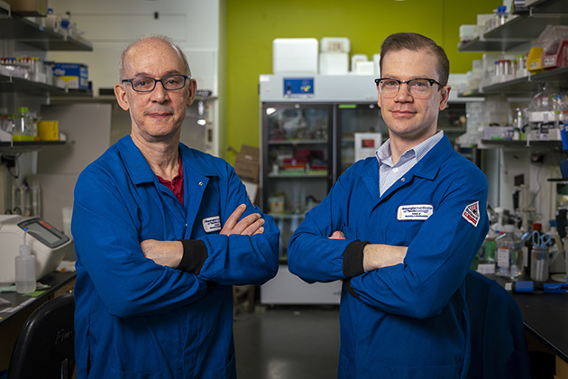 Two people wearing blue lab coats stand with arms crossed in a laboratory filled with shelves, equipment, and a large refrigerator in the background.
