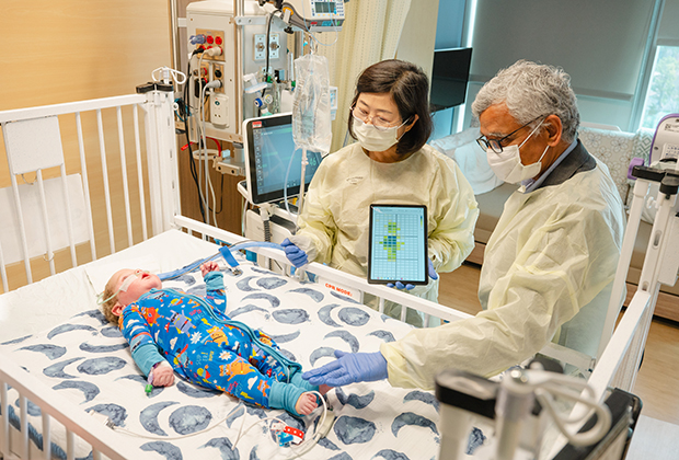 Two adults wearing protective gowns and gloves stand beside a hospital crib, using a tablet device while examining an infant lying on the mattress as medical equipment and monitors surround the crib.

