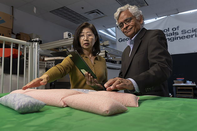 Two adults in a laboratory examine soft sensor‑embedded crib materials on a padded surface, with one person holding a tablet displaying data.