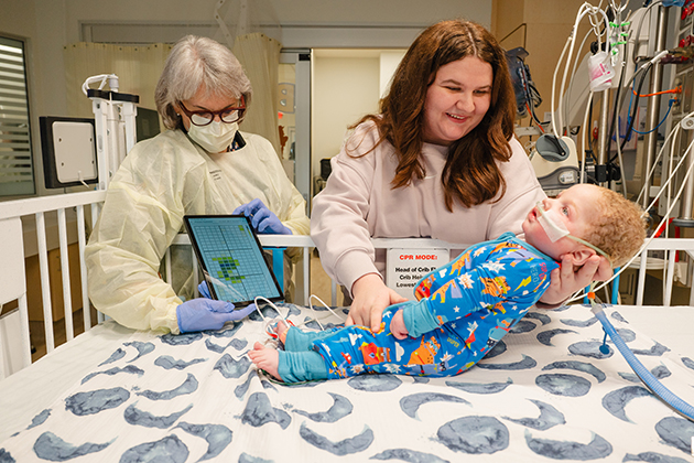 One adult wearing a protective gown and gloves stands beside a hospital crib holding a tablet displaying data, while another adult supports an infant positioned on the crib mattress amid nearby medical equipment.