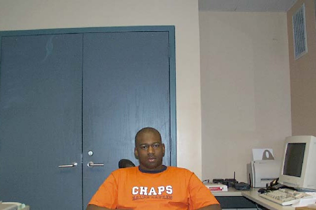 A person in an orange “CHAPS” T‑shirt sits at a desk in a small office with a closed blue door and an older desktop computer nearby.
