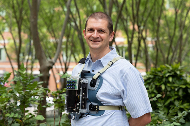 man wearing a light blue shirt standing in front of several trees with a device strapped to his chest