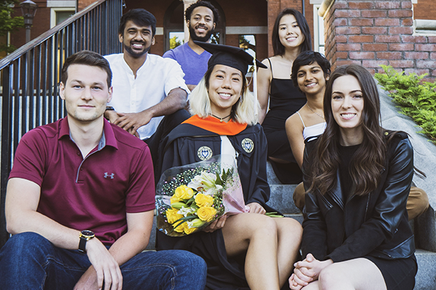 A group of people sitting on outdoor steps in front of a brick building, with one person wearing a graduation cap and gown holding a bouquet of yellow flowers.