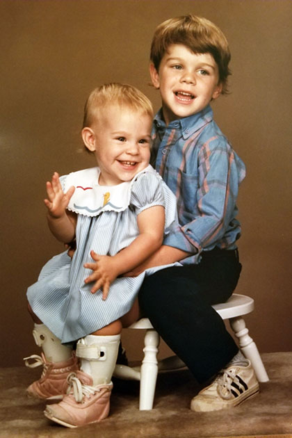 Two young children sitting on a small white stool, with the older child wearing a striped shirt and sneakers, and the younger child in a light blue dress and orthopedic braces on both legs.