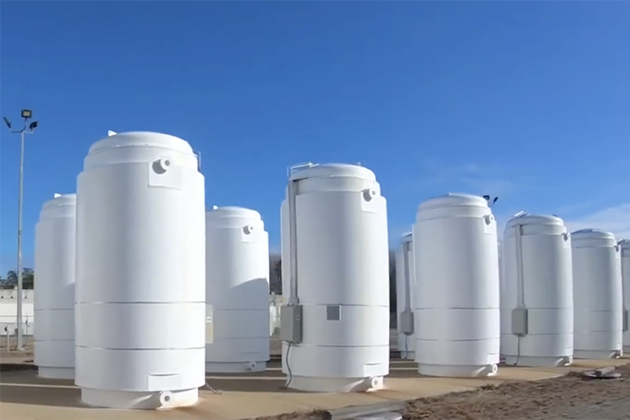 Several large, white cylindrical dry storage casks arranged outdoors on a concrete pad under a clear blue sky.