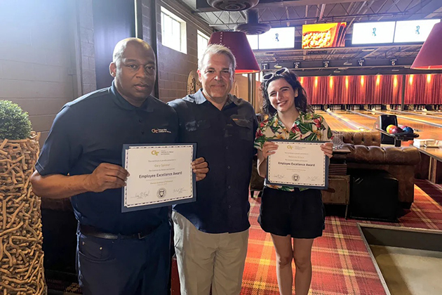 Three people stand in a bowling alley holding “Employee Excellence Award” certificates and smiling in front of the lanes and lounge seating.