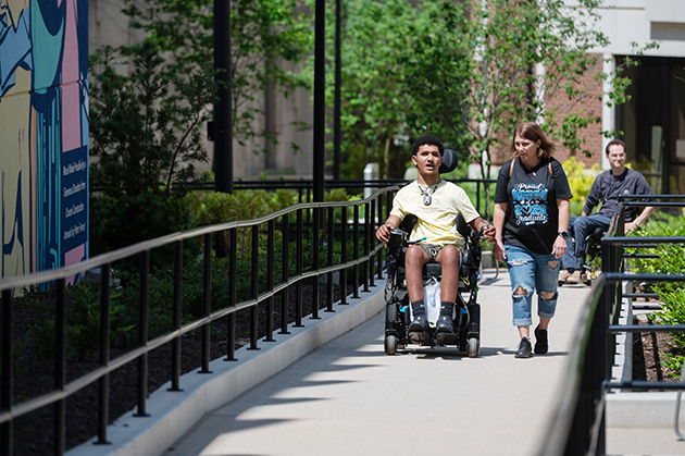 Person using a motorized wheelchair travels along an outdoor ramp with another individual walking beside them, near a building and landscaped area.