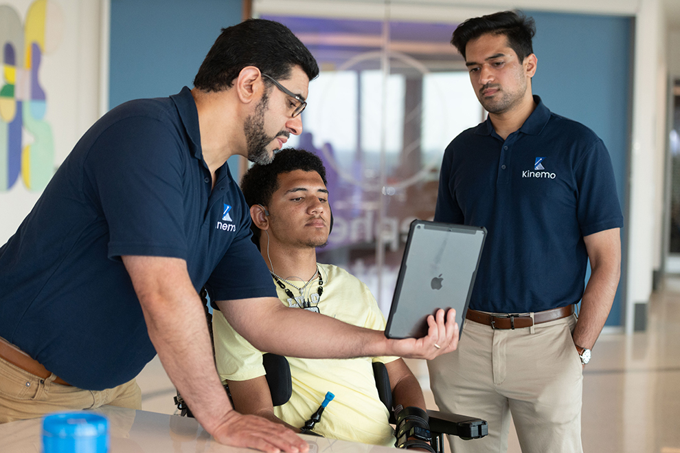 Two Kinemo staff members show a tablet to a young wheelchair user wearing assistive technology during a collaborative office session.