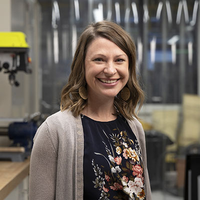 Woman in a workshop environment with industrial equipment and tools in the background, wearing a floral-patterned blouse and light knit cardigan, representing a modern manufacturing or maker space setting.