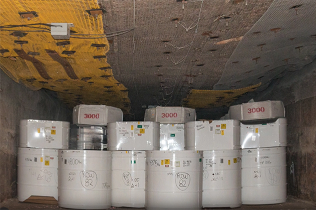 Underground storage area with multiple white cylindrical barrels labeled with numbers and markings, used for storing low-level radioactive waste, beneath a reinforced ceiling with mesh and yellow panels.