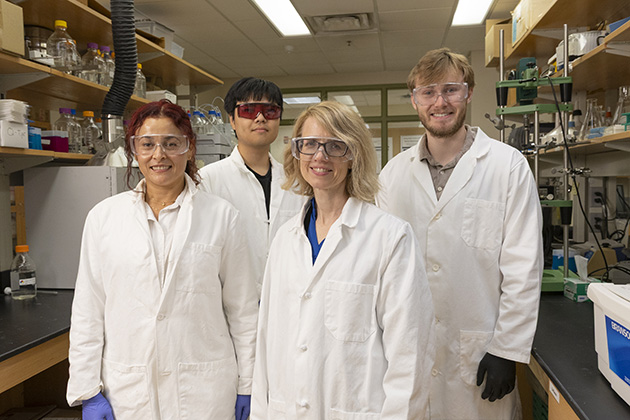Four individuals wearing white lab coats stand together in a laboratory setting. 