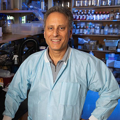 Researcher in a lab wearing a blue coat, standing in a laboratory with shelves of bottles and equipment in the background.