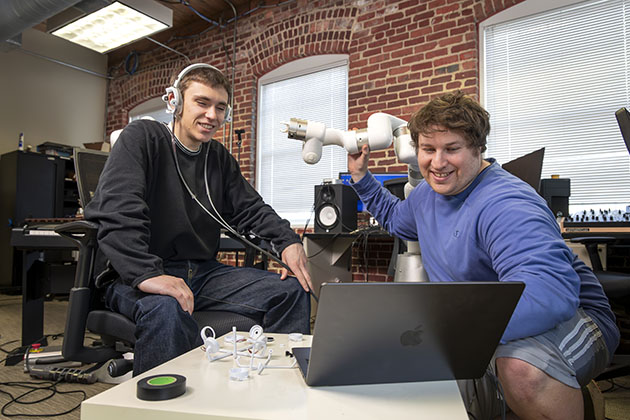 Two individuals working at a laptop in a lab with a robotic arm and small components on a table surrounded by brick walls and equipment.