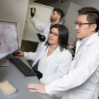 Several people wearing white lab coats gather around a computer monitor displaying a magnified scientific image in a laboratory.
