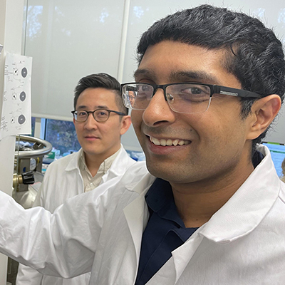 People wearing white lab coats stand near laboratory equipment, examining controls and instrumentation.

