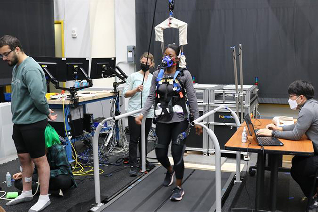 Person walking on a treadmill wearing a harness and sensors, with researchers monitoring equipment and computers in a lab setting.