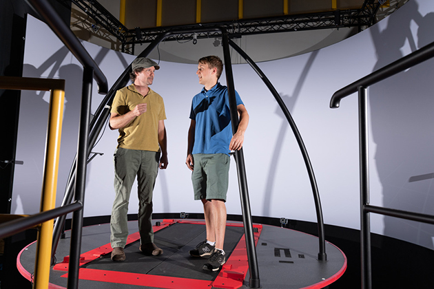 Two people standing on a circular platform with metal support bars in a research or testing environment, surrounded by curved walls and overhead framework.