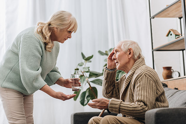 Older adult offering a glass of water and medication to another older adult seated on a couch in a living room with plants and shelves in the background.