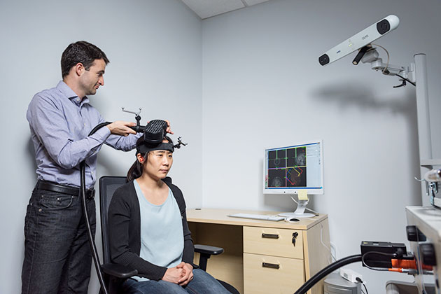 Researcher adjusting a device on another person’s head in a lab, with a computer displaying brain imaging data and a mounted camera in the background.