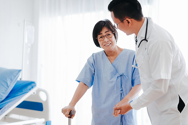 Healthcare professional assisting a patient in a hospital room, with the patient using a cane and wearing a hospital gown and a bed and medical equipment visible in the background.