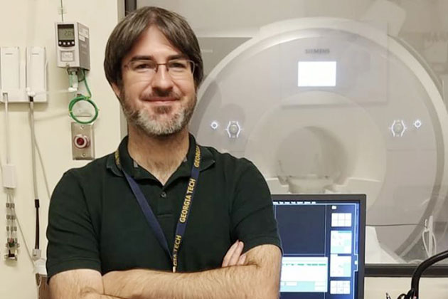 Individual standing in front of an MRI machine with arms crossed, wearing a lanyard, and a computer monitor visible nearby.