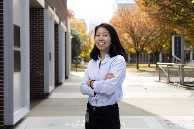 Woman standing outdoors on a paved walkway with arms crossed, wearing a light blue button-up shirt and dark pants. The background shows brick buildings, trees with autumn foliage, and a metal railing.