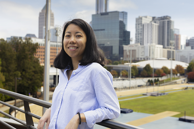 Woman wearing a light blue button-up shirt leaning on a metal railing with a city skyline in the background. The scene includes tall modern buildings, a sports field, and trees with autumn foliage under a partly cloudy sky.