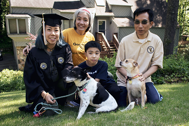 Family of four people sitting on a grassy lawn in front of a house, with two dogs in the foreground. One person is wearing a graduation cap and gown, another is wearing a yellow Georgia Tech shirt, and the others are dressed casually.