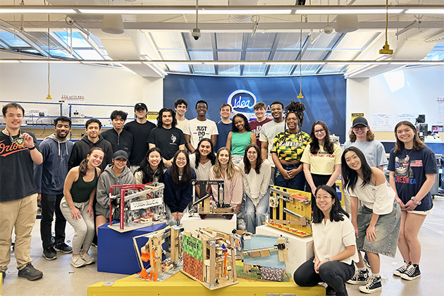 Group of students posing together in a bright workshop or makerspace, standing and sitting around colorful mechanical projects displayed on tables. The background features a large blue wall with the word 'Idea' and shelves with tools and equipment.