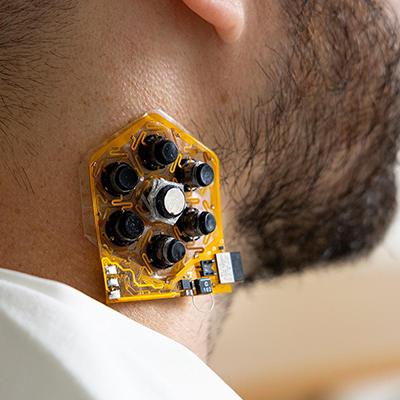 A close-up view of a small hexagonal electronic sensor attached to the side of a person’s neck, featuring multiple black components and flexible circuitry.